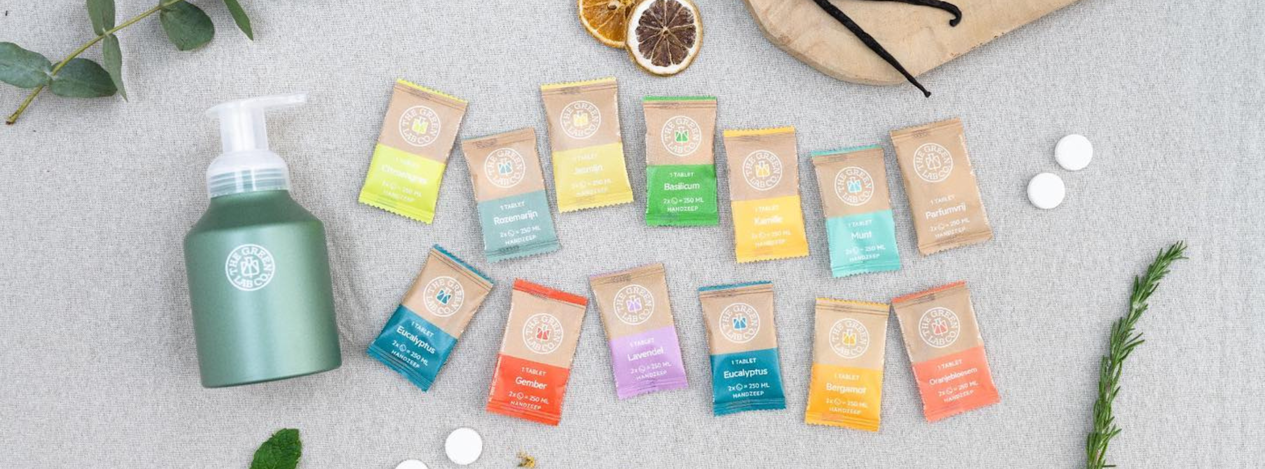 Flera färgglada förpackningar med handtvålstabletter från The Green Lab Co. ligger på tyg, bredvid en grön tvålpump.