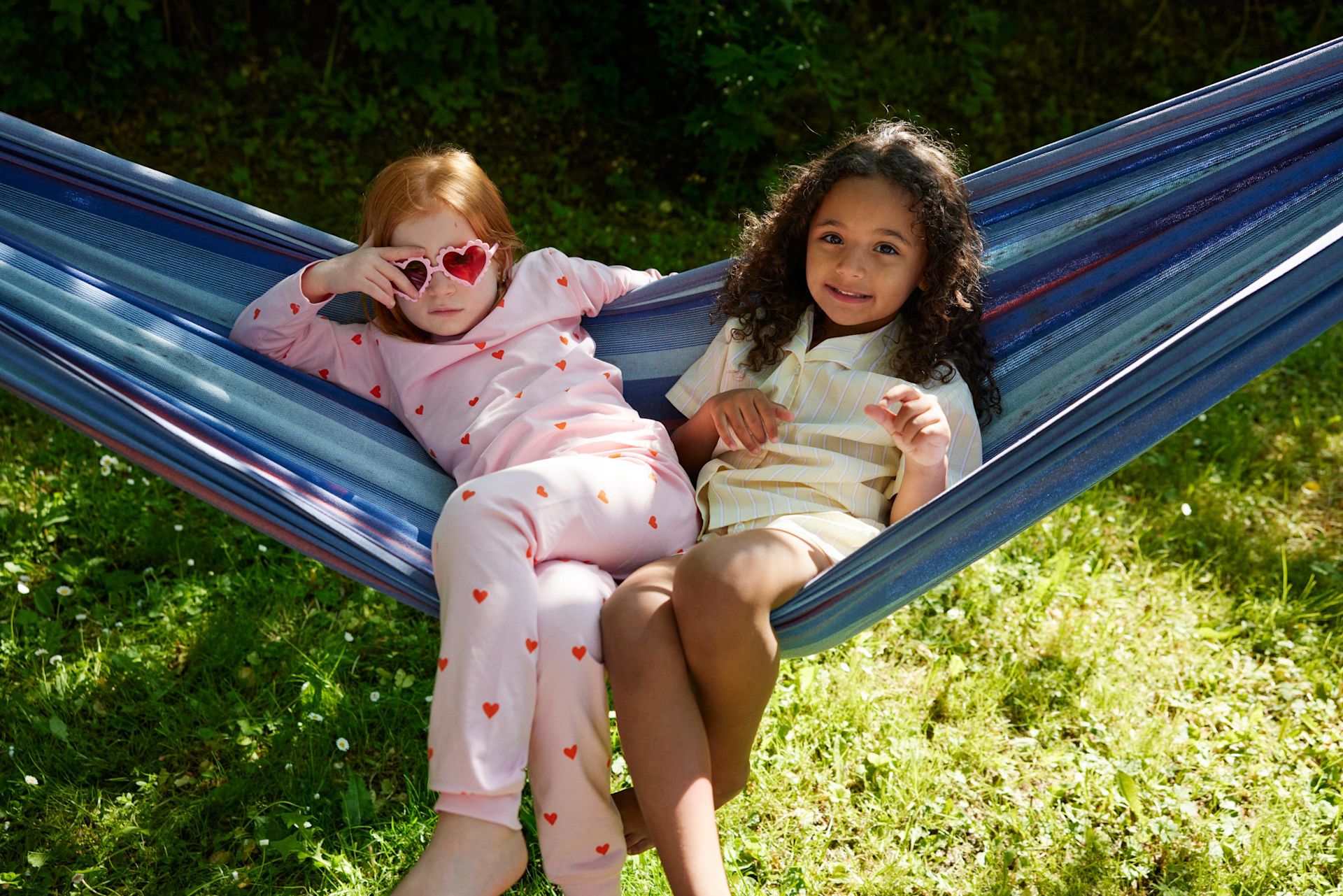 Två flickor i 5-6-årsåldern sitter tillsammans i en hängmatta och tittar mot kameran. Det är sommar och sol