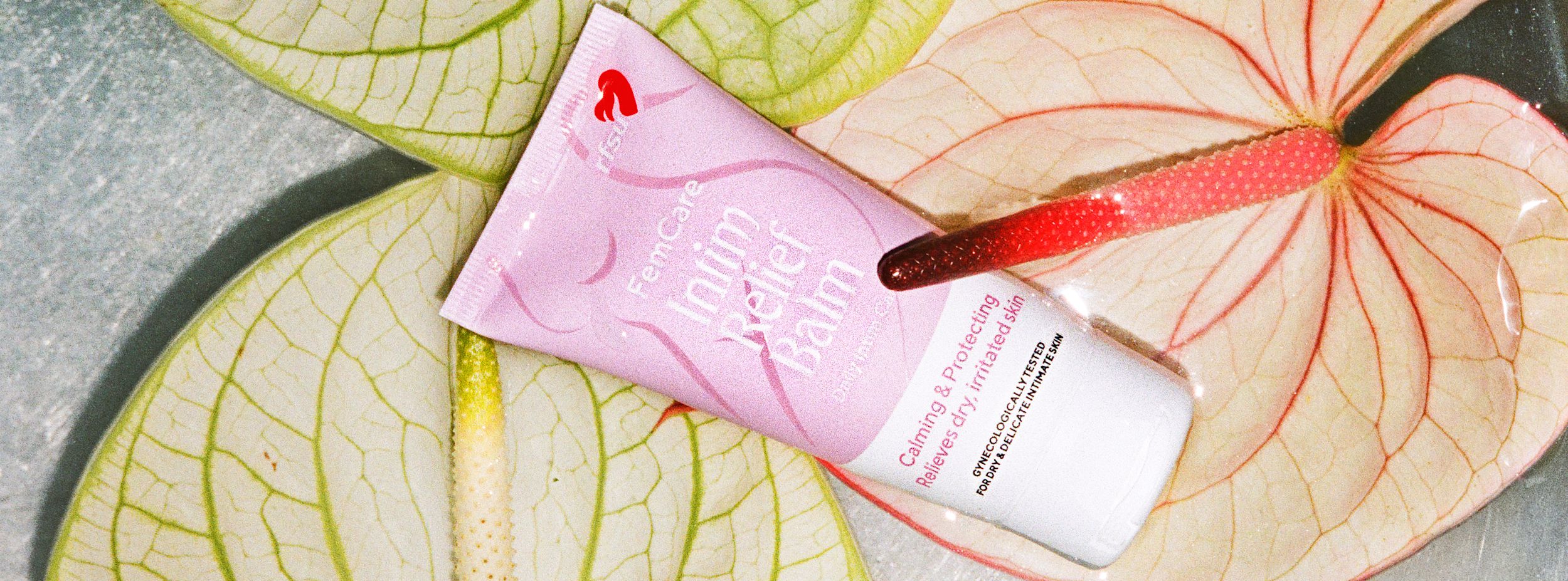 En rosa tub med FemiCare Intim Relief Balm ligger på stora, ljusa blad med röda ådror.