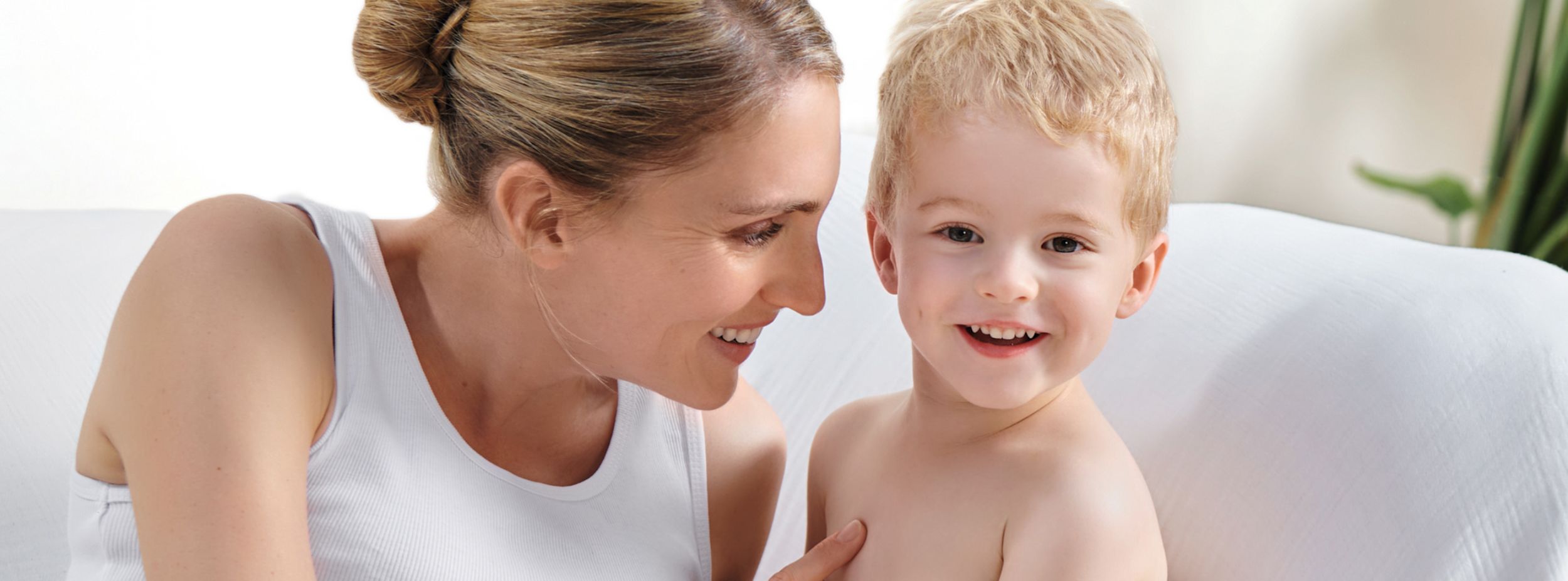 Leende kvinna sitter nära ett glatt litet barn på en vit soffa i ett ljust rum.