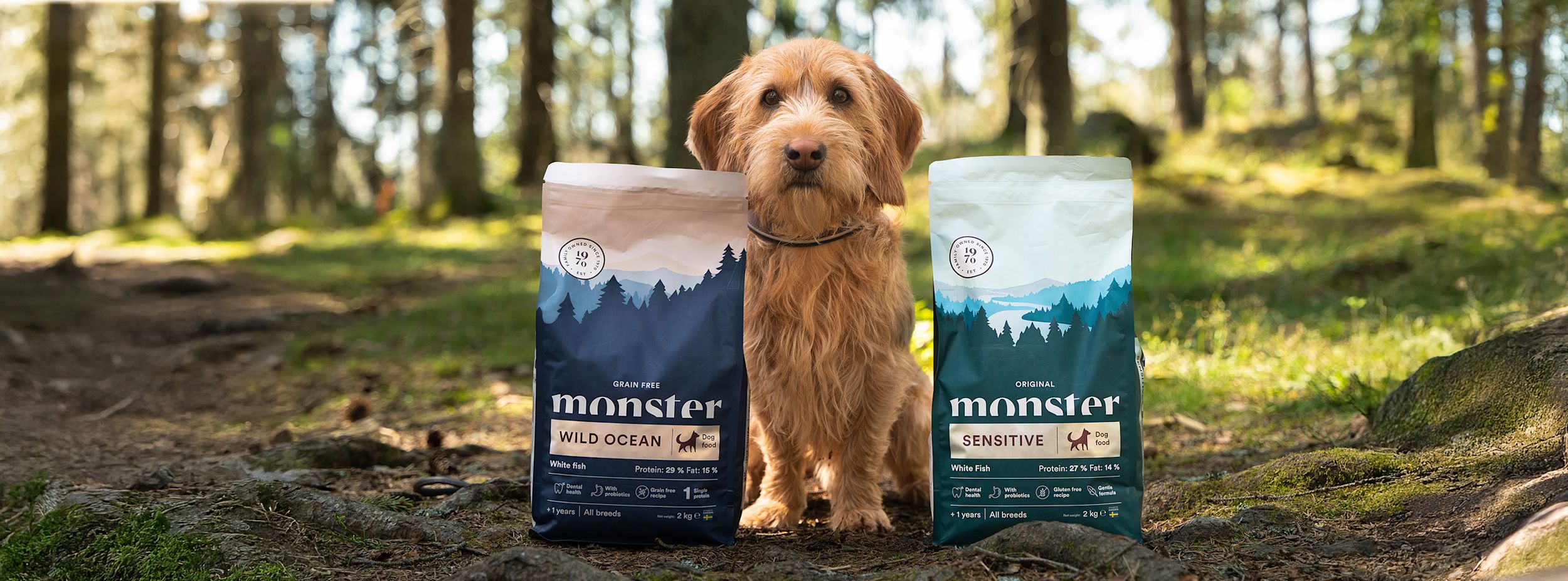 Hund sitter i skogen mellan två påsar Monster hundmat, Wild Ocean och Sensitive, båda med blå och gröna förpackningar.