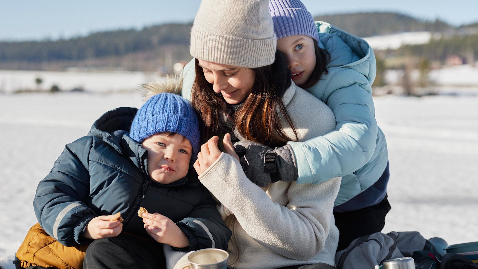 Mamma sitter med två barn utomhus i snön, iklädda mössor och varma jackor och fikar tillsammans.