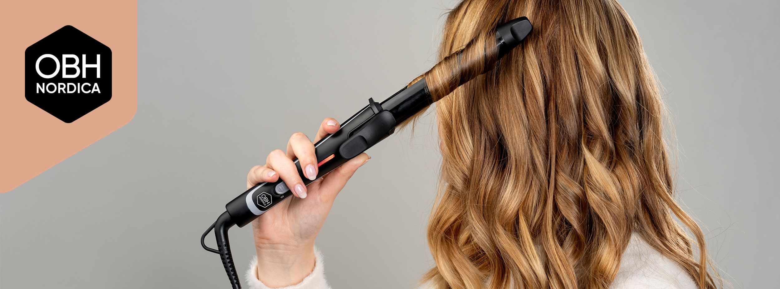 Kvinna lockar sitt blonda hår med en svart OBH Nordica-locktång, logotyp syns till vänster mot grå bakgrund.