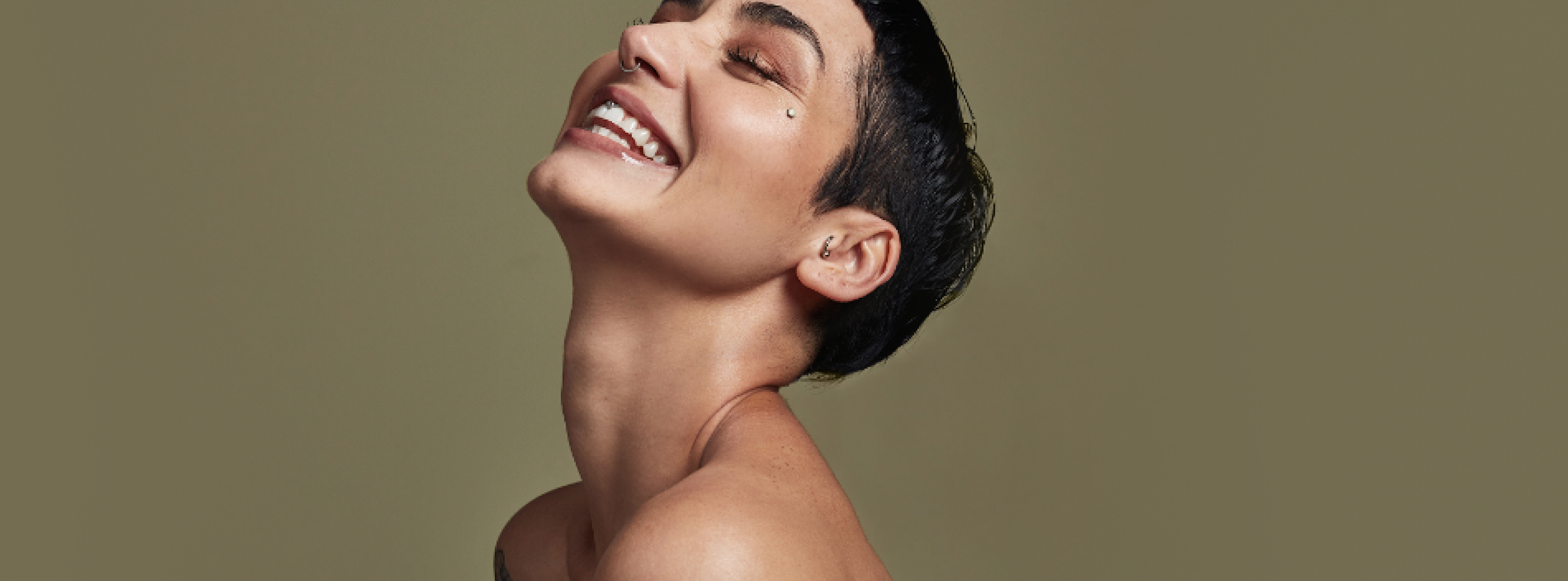 Person med kort mörkt hår skrattar och lutar huvudet bakåt mot en enfärgad bakgrund.