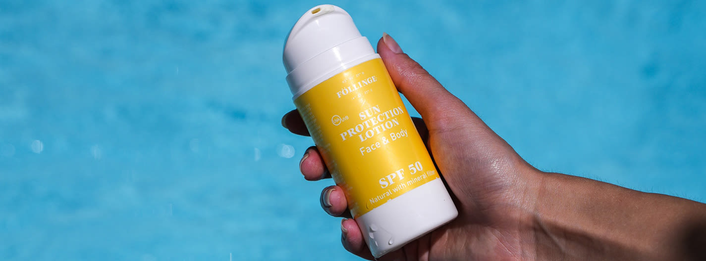 En hand håller en flaska Föllinge Sun Protection Lotion SPF 50 framför en blå bakgrund, troligen vid en pool.