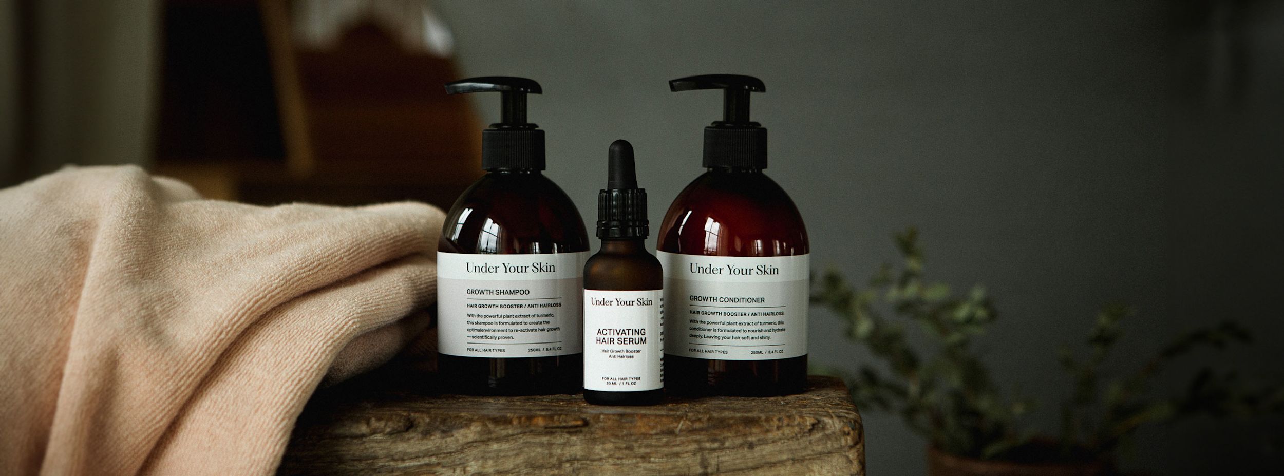 Tre flaskor Under Your Skin hårvårdsprodukter står på en träyta bredvid en beige handduk och en grön växt i bakgrunden.