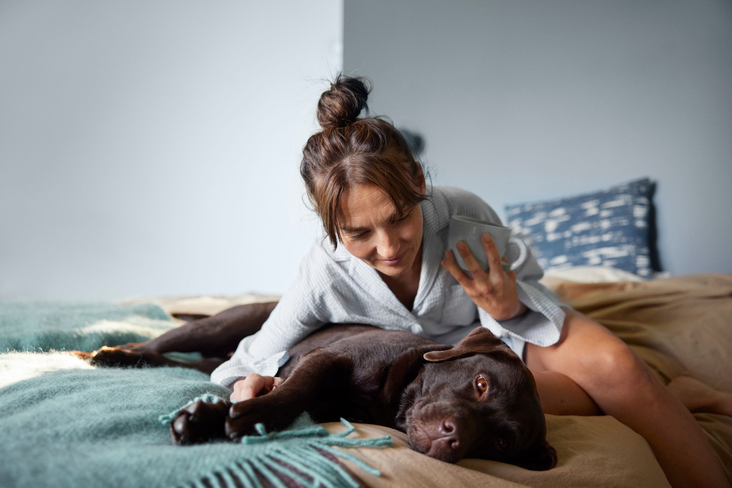 Kvinna sitter i sängen tillsammans med en hund som ligger ner. Hon lutar sig fram och klappar hunden.