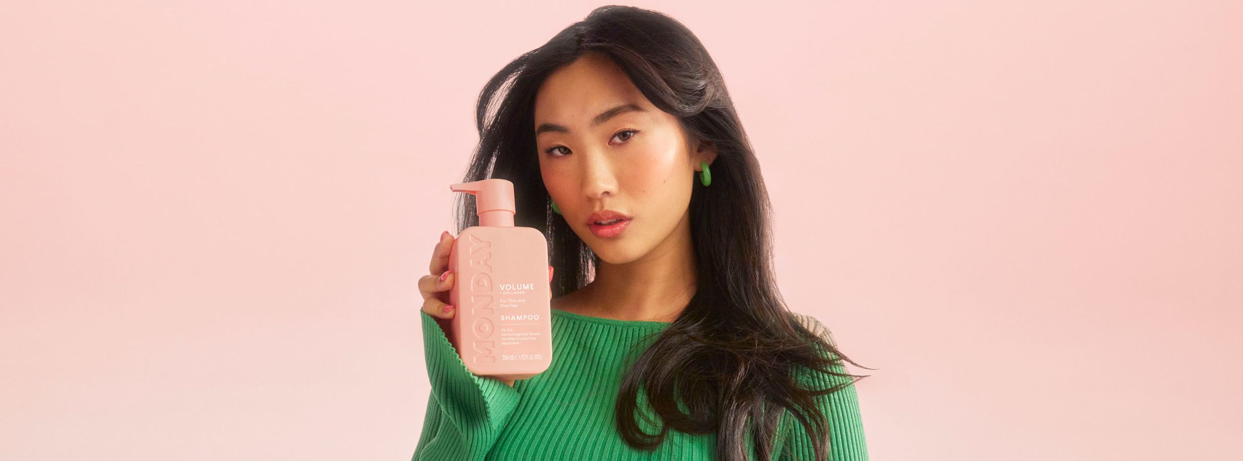 Kvinna i grön tröja håller upp en rosa flaska med texten "MONDAY Volume + Collagen Shampoo" mot en ljusrosa bakgrund.