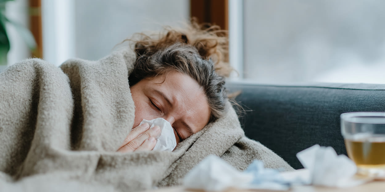Kvinna ligger nedbäddad i soffan med filten högt uppdragen. Hon snyter sig och ser väldigt hängig ut.
