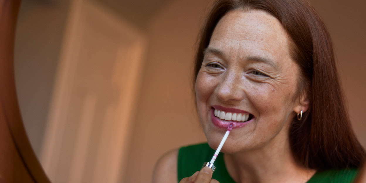 Kvinna i medelåldern med ett stort varmt leende står framför spegeln och applicerar läppglans. Hon har mörkt långt hår och och man kan se ljusa pigmentfläckar i hennes hud i ansiktet.