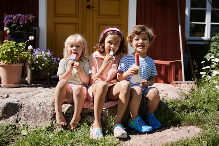 Tre barn sitter utomhus på sommaren i solskenet och äter glass tillsammans. 