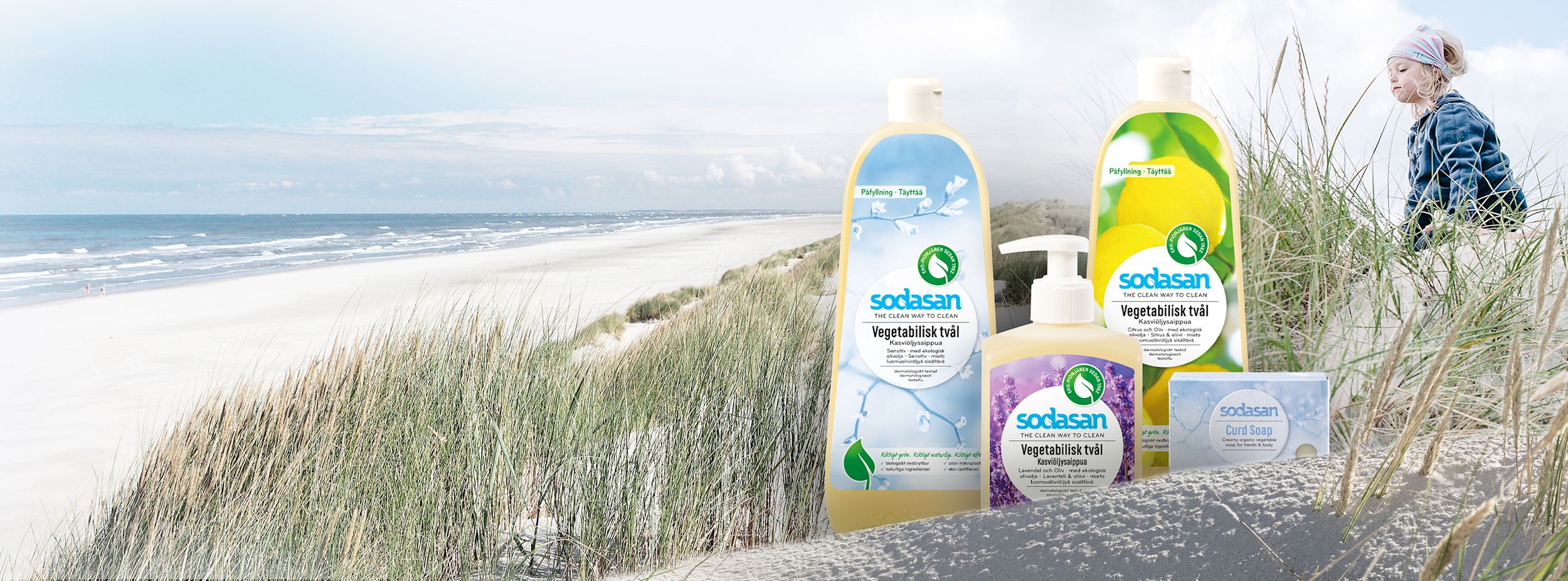 Fyra flaskor Sodasan vegetabilisk tvål står på en sandstrand med gräs, hav i bakgrunden och ett barn som leker i sanddynerna.
