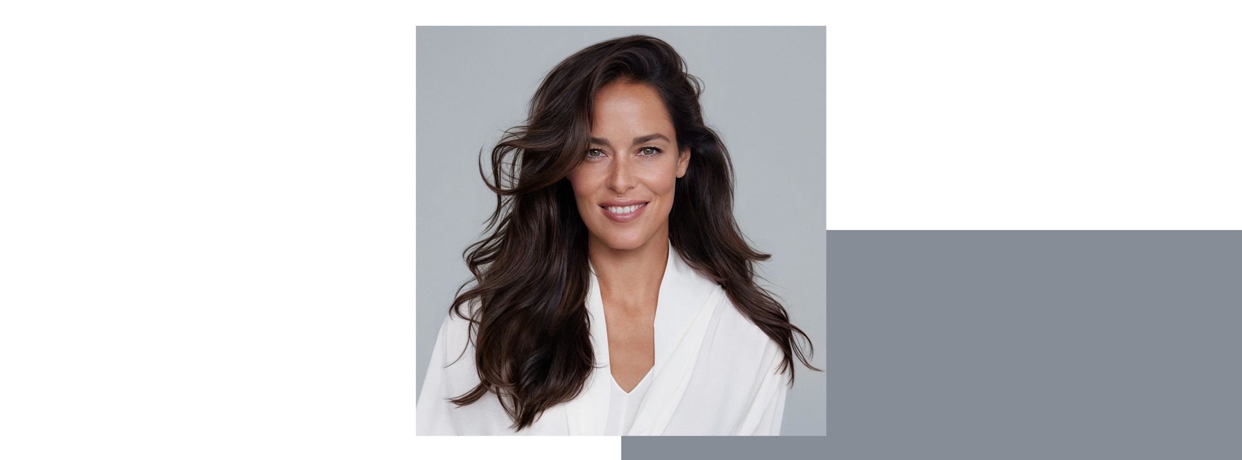 Kvinna med långt, mörkt vågigt hår ler mot kameran, klädd i vit skjorta mot ljusgrå bakgrund.