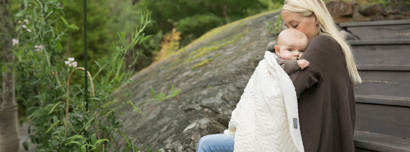 Kvinna sitter utomhus på trappa och håller en bebis inlindad i vit filt, omgiven av grönska och stenar.