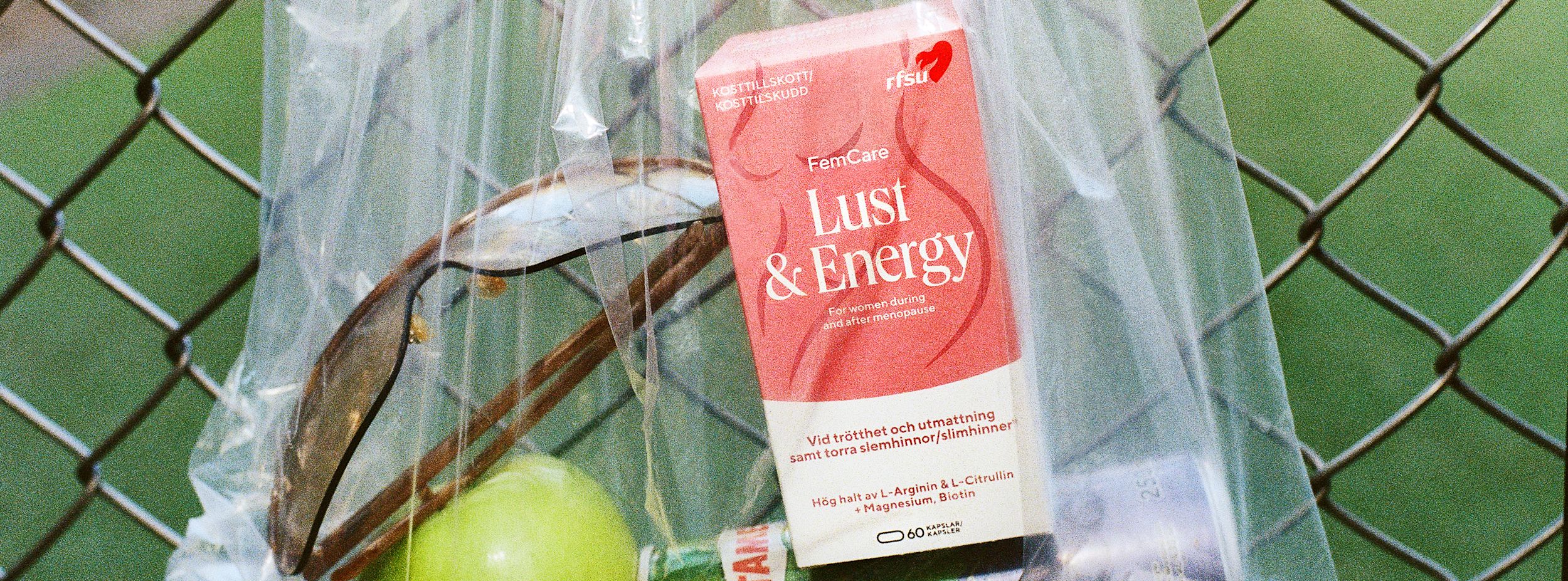 Kosttillskottet "Lust & Energy" från RFSU ligger i en plastpåse mot ett stängsel, tillsammans med ett grönt äpple och solglasögon.