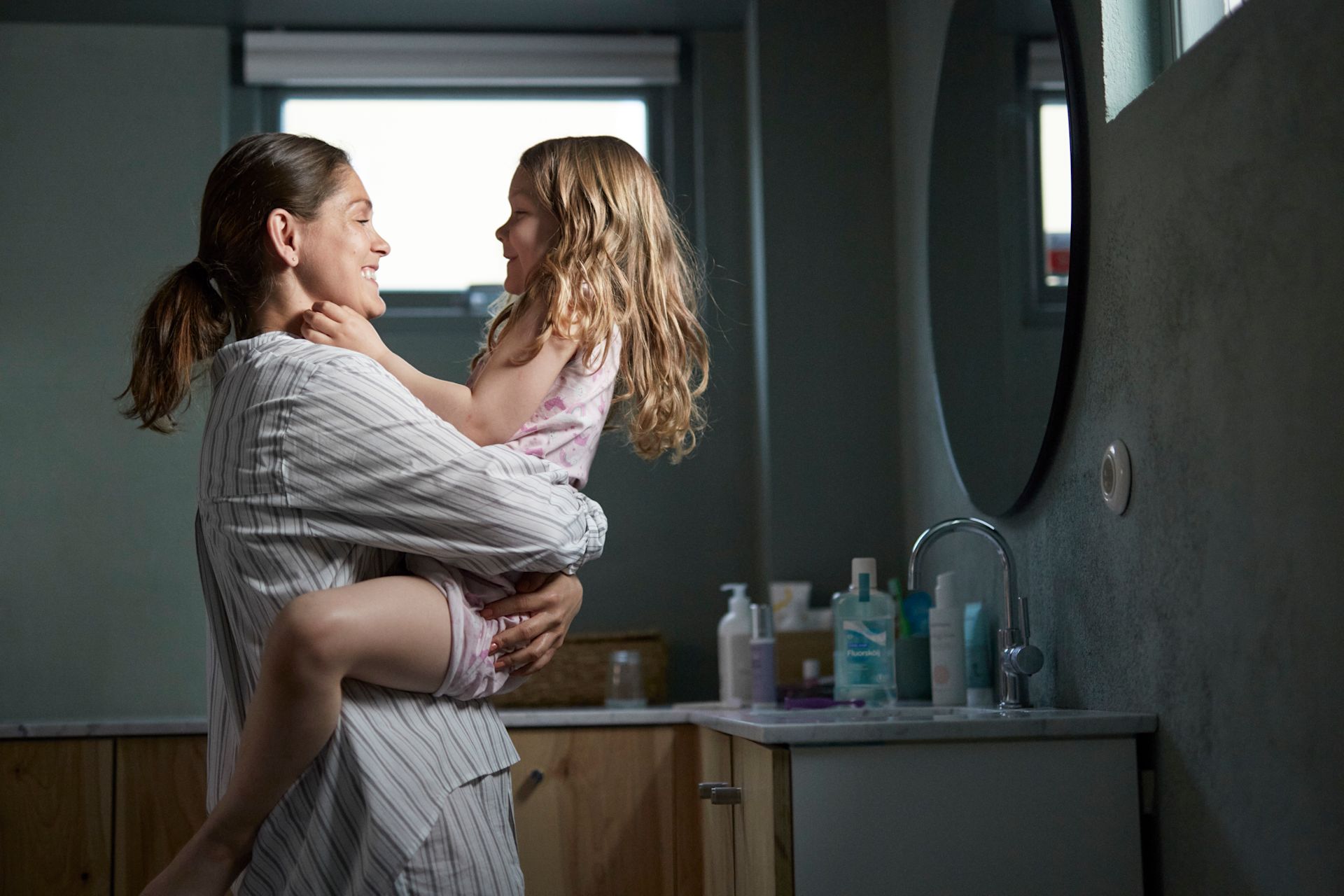 Mamma och dotter är i badrummet. Mamma håller sin dotter i famnen och båda skrattar.