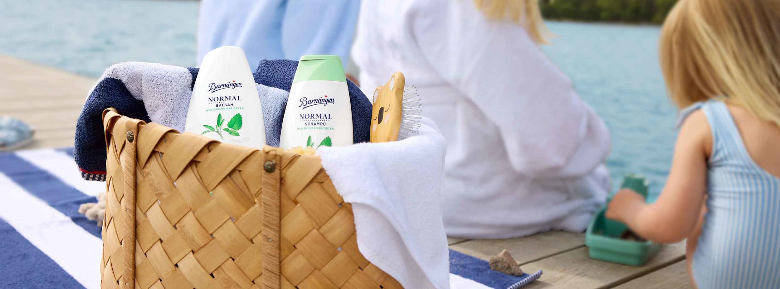 Korg med Barnängen Normal balsam och schampo, borste och handdukar på brygga vid sjö, barn leker i bakgrunden.