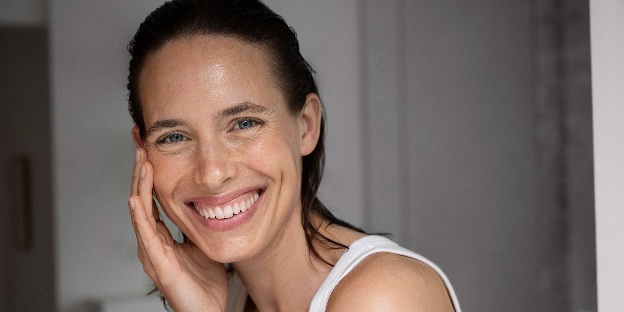 Kvinna tittar in i kameran och ler med ett stort leende. Hon håller några fingrar lätt på ena kinden.