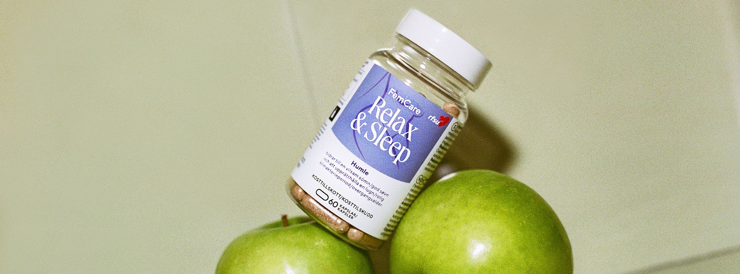 Burk med kosttillskottet FemCare Relax & Sleep balanserar på två gröna äpplen mot ljus bakgrund.