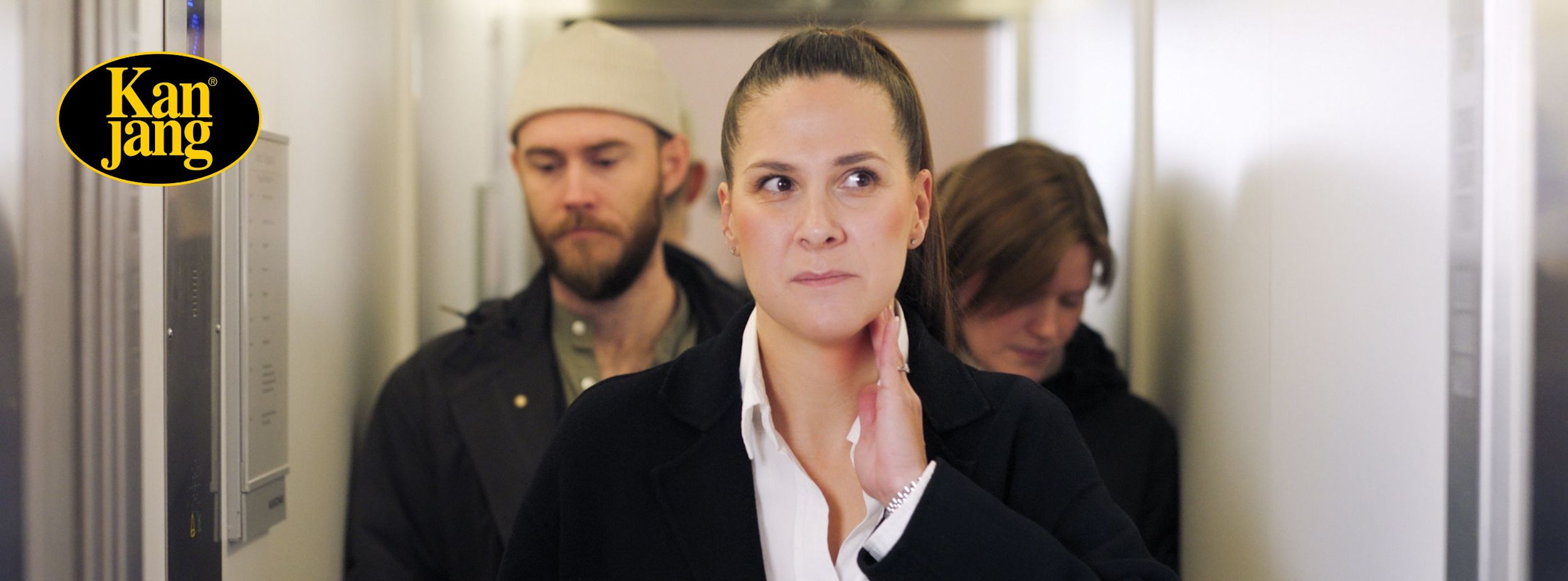 Kvinna rör vid sin hals och ser bekymrad ut i en hiss med två andra personer, Kan Jang-logotyp syns till vänster.
