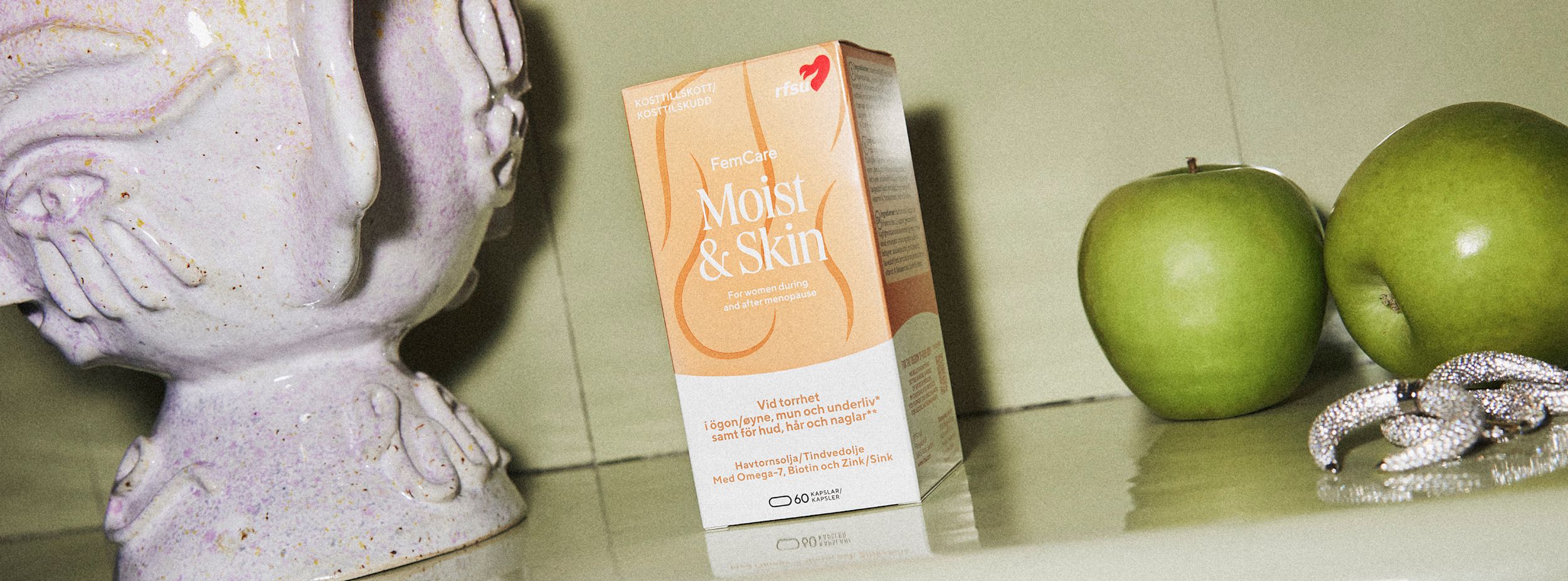 En förpackning FemCare Moist & Skin kosttillskott står på ett bord bredvid två gröna äpplen, smycken och en ljus keramikvas.