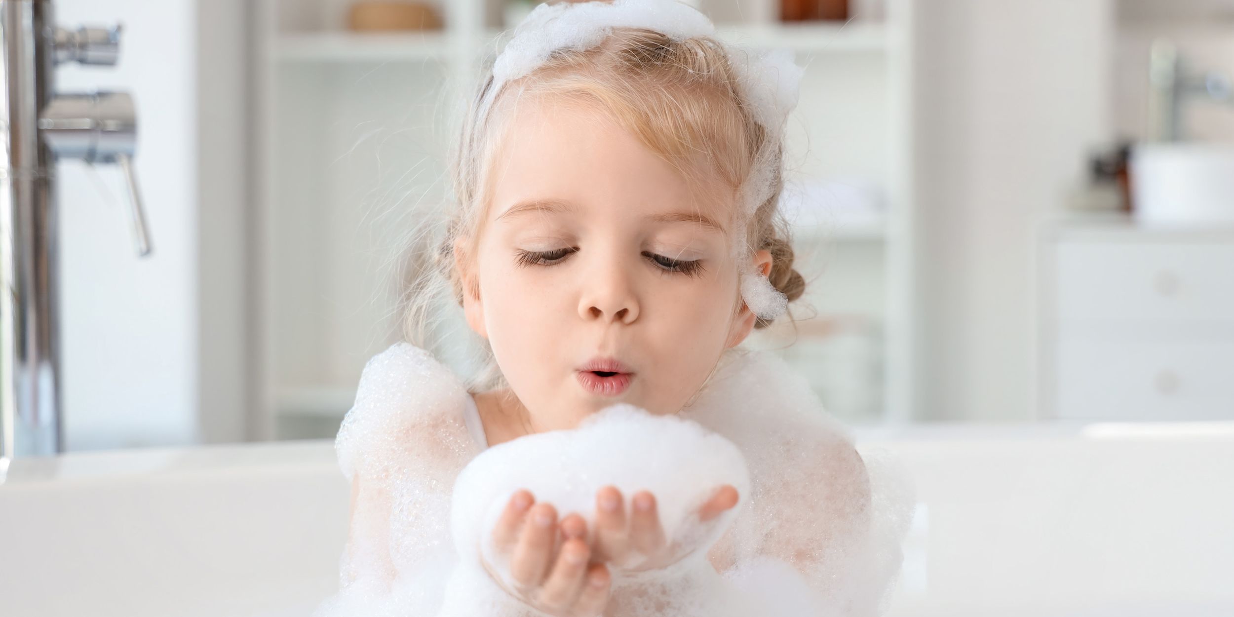 En flicka sitter i badkaret och håller badskum i handen som hon blåser på.