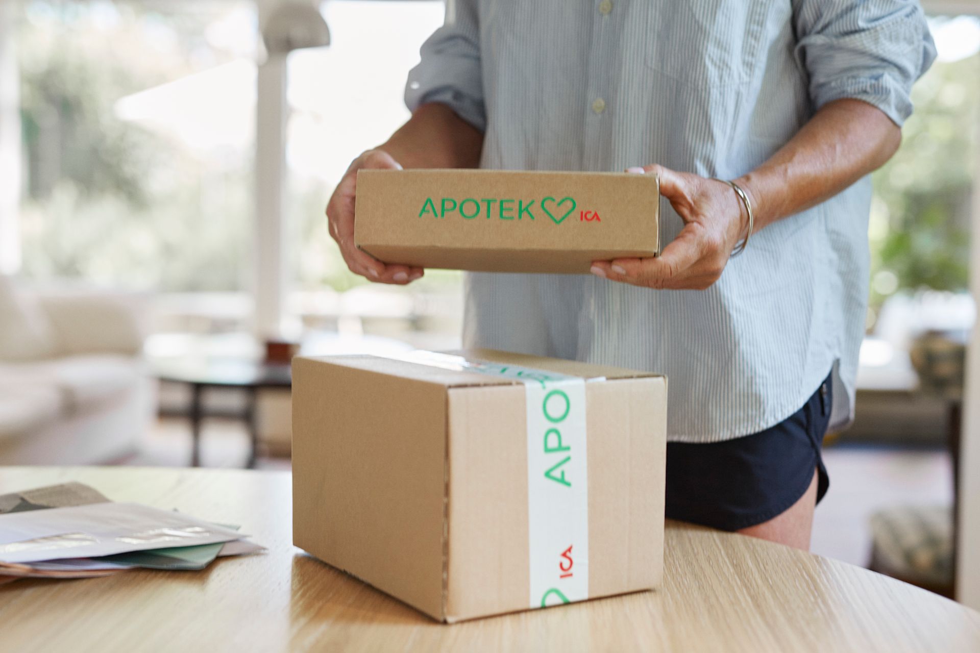 Kvinna står framför ett bord med flera brev liggandes i en hög och två paket från Apotek Hjärtat, och håller upp ett i handen.