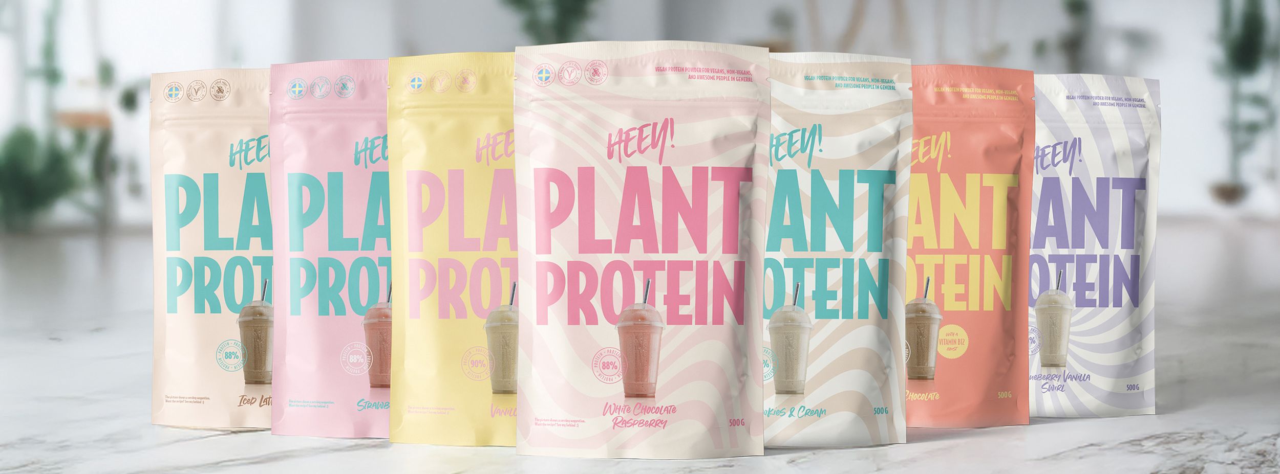 Sju färgglada påsar med texten "HEEY! PLANT PROTEIN" står uppradade på ett ljust köksbord, varje påse visar olika smaker och färger.