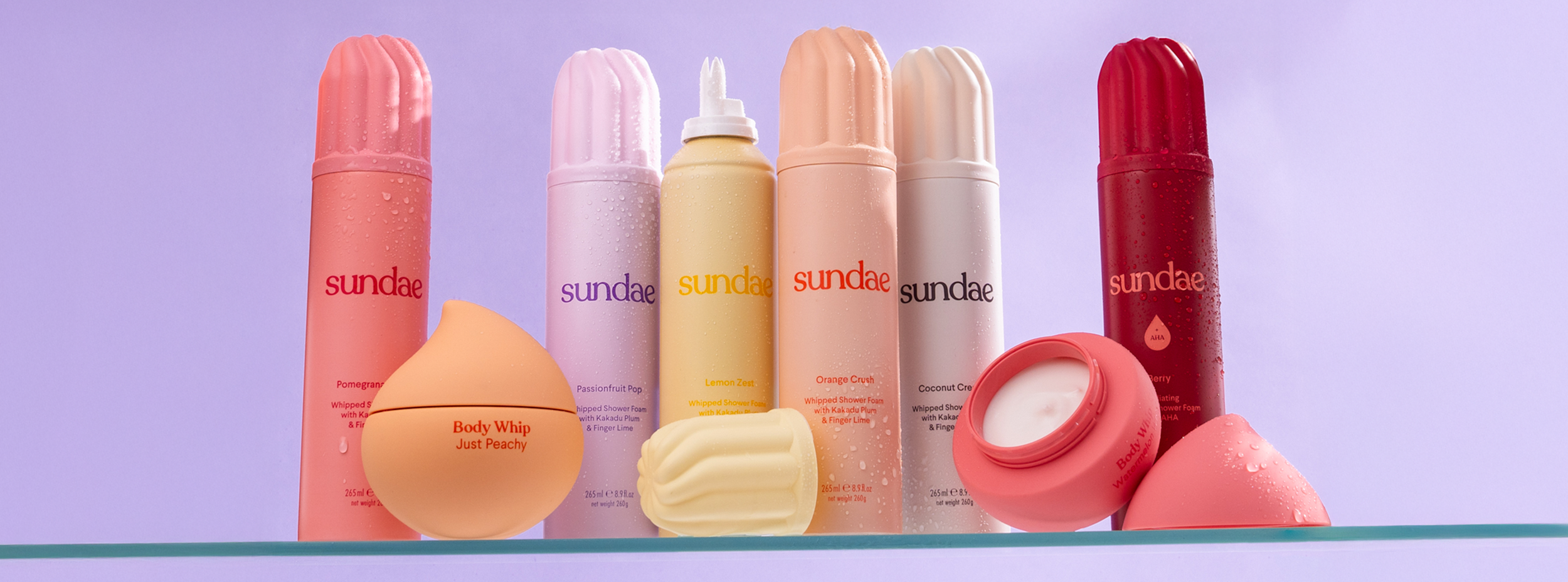 Flera flaskor och burkar av Sundae kroppsvårdsprodukter står uppradade på en glasyta mot en lila bakgrund.