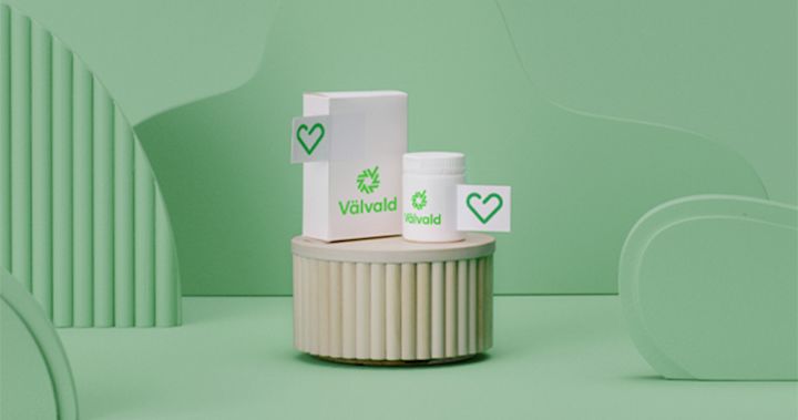 På en cylinder i trä står två vita förpackningar märkta med Apotek Hjärtats symbol, en ask och en burk. Tryckt på förpackningarna står det med grönt ordet välvald och dess symbol.