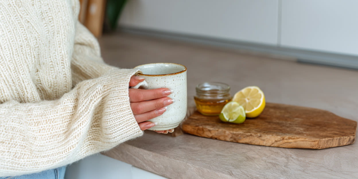 Bild på en köksbänk där citron, lime och honung är framplockad. Vid sidan av syns en kvinna som håller en tekopp 