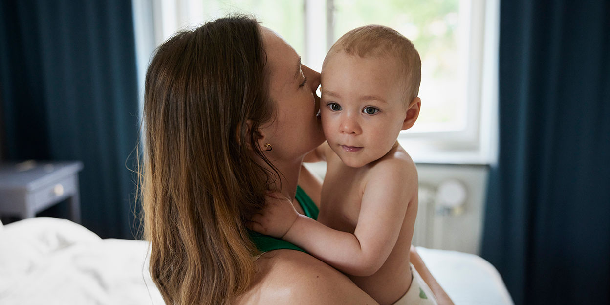 Mamma och litet barn i sovrumsmiljö. Mamman håller barnet i sin famn.