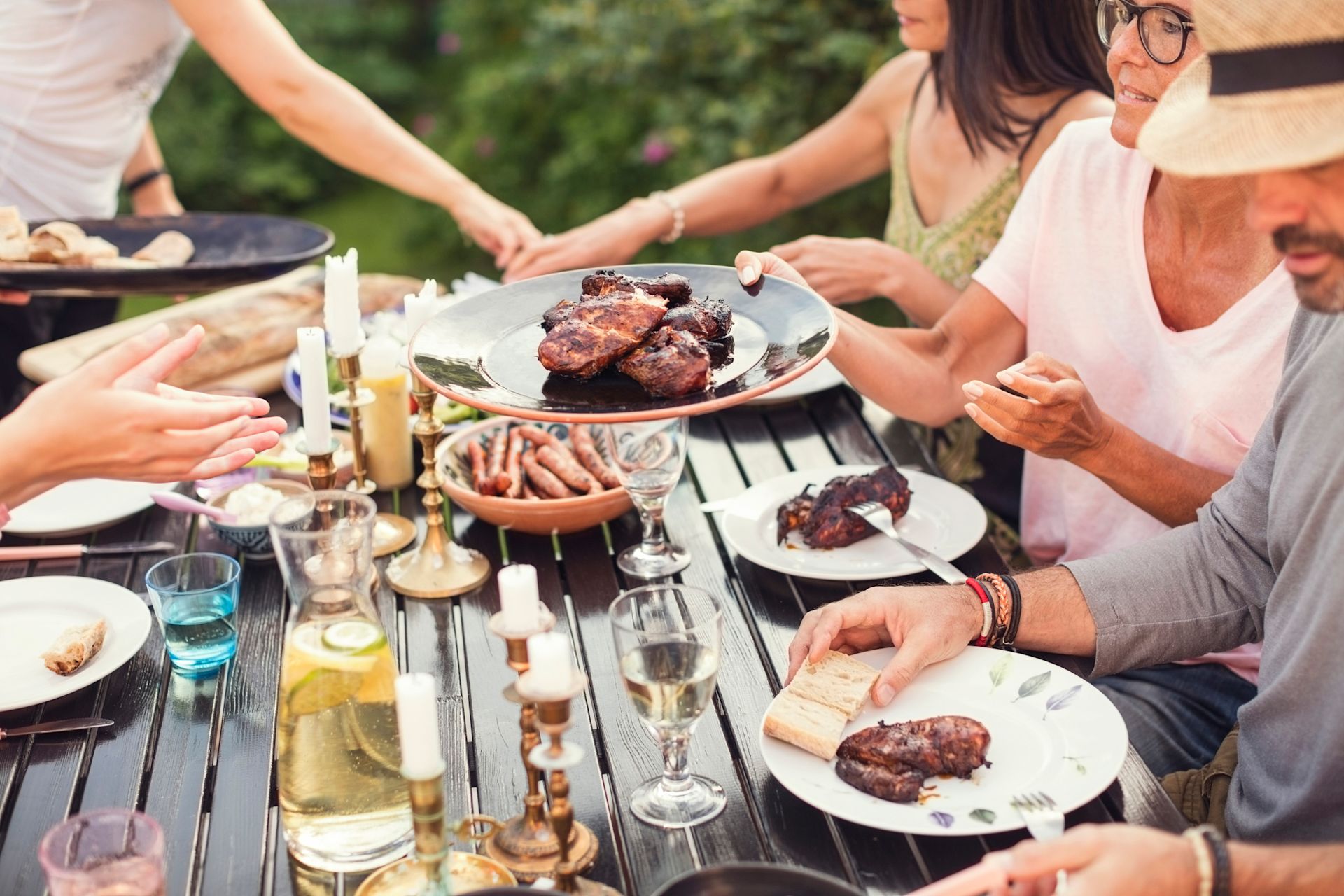 Människor sitter utomhus kring ett bord och äter grillat kött till middag en sommarkväll.