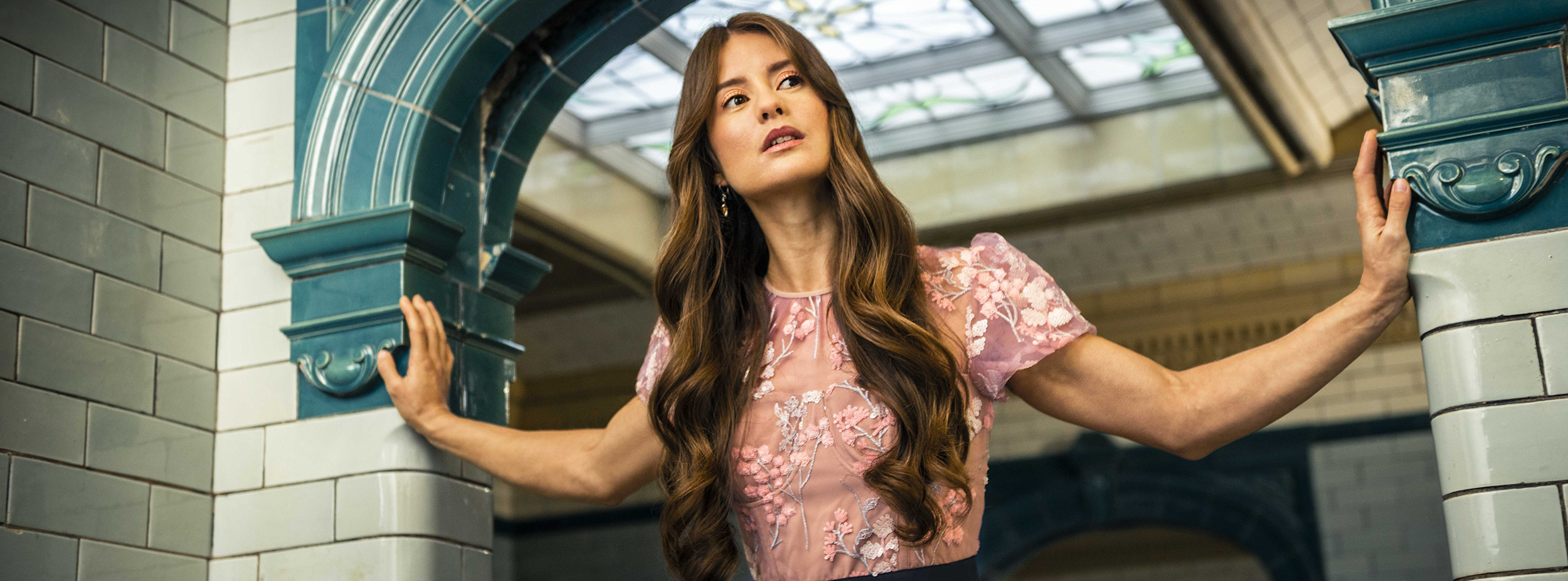 Kvinna med långt brunt hår håller i två pelare, bär en rosa blus med blommönster, står i ett rum med kaklade väggar och glastak.