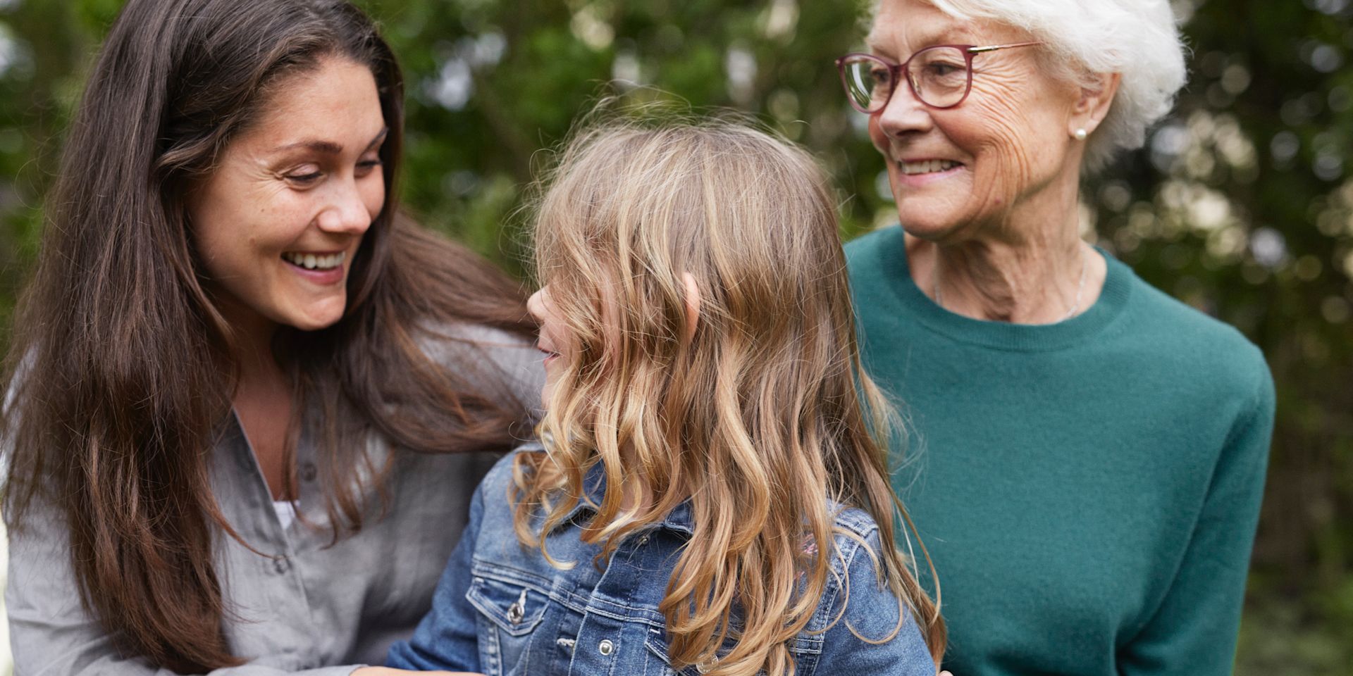Mormor, mamma och dotter sitter utomhus tillsammans och kramas och skrattar.